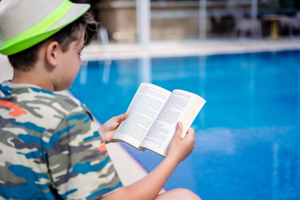 Boy in a cap reading a book beside a swimming pool