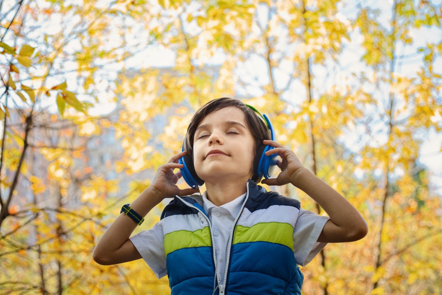 Smiling boy listening to headphones with his eyes closed among yellow autumn leaves