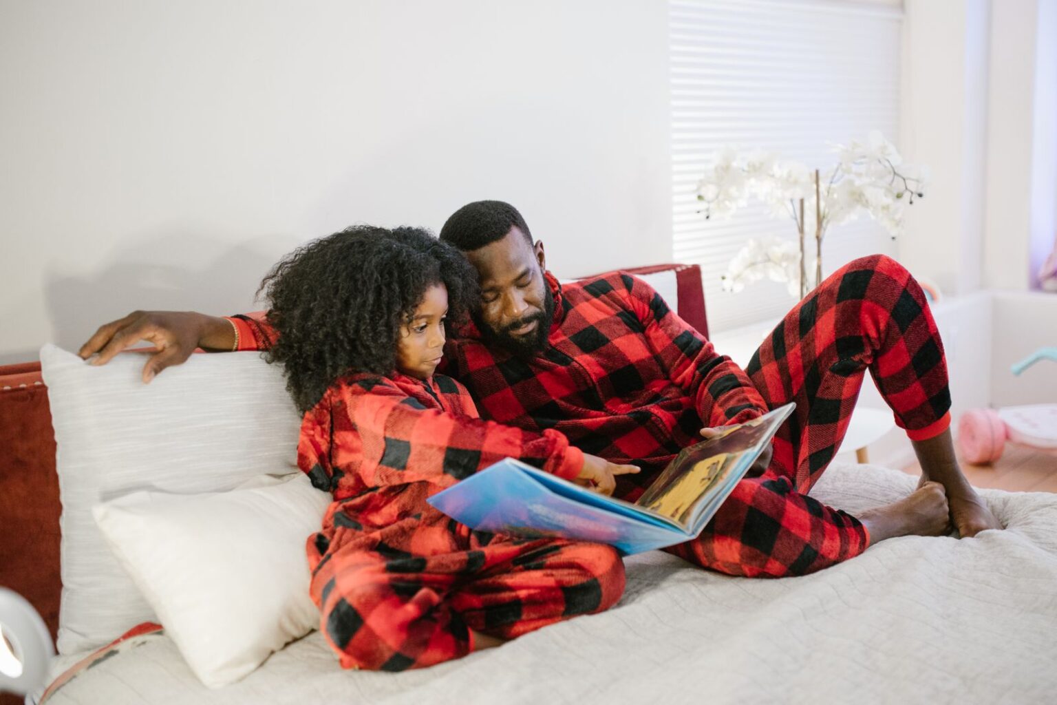 Father and daughter in matching red plaid pajamas reading a picture book on the bed