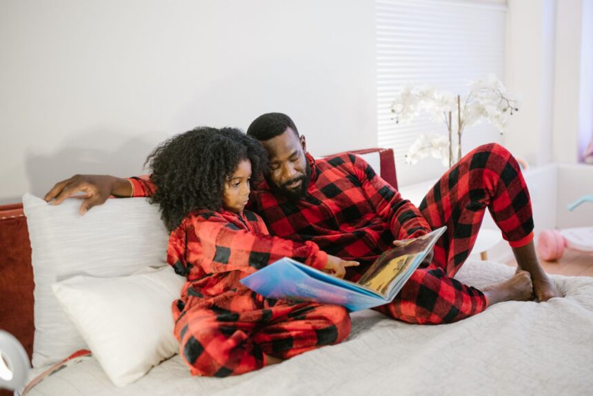 Father and daughter in matching red plaid pajamas reading a picture book on the bed