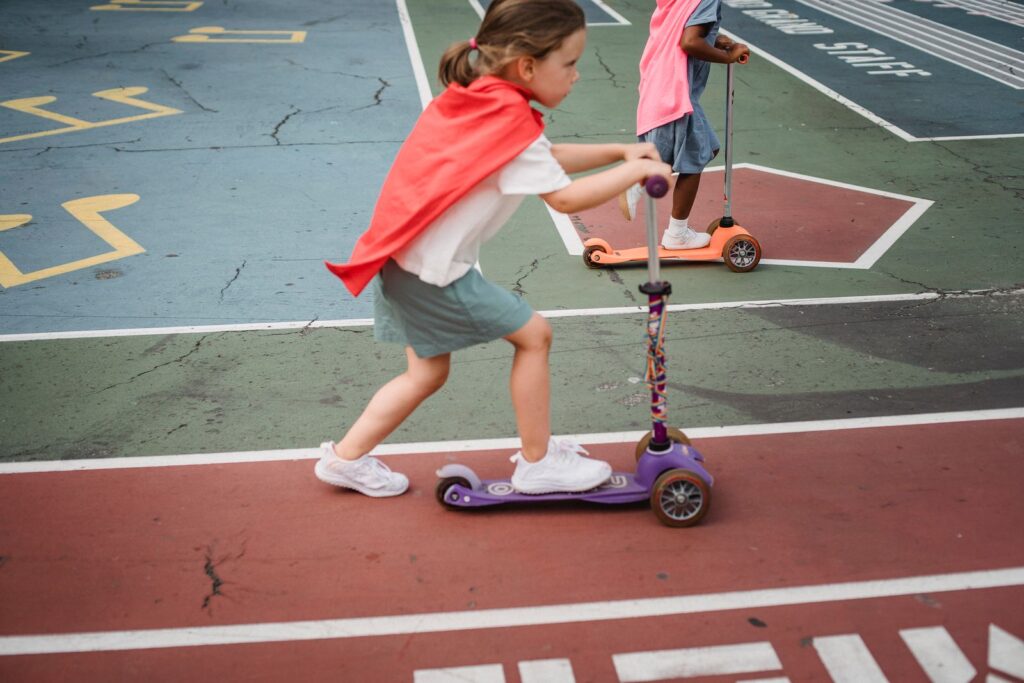 Girls in capes riding three-wheel kick scooters on a playground court