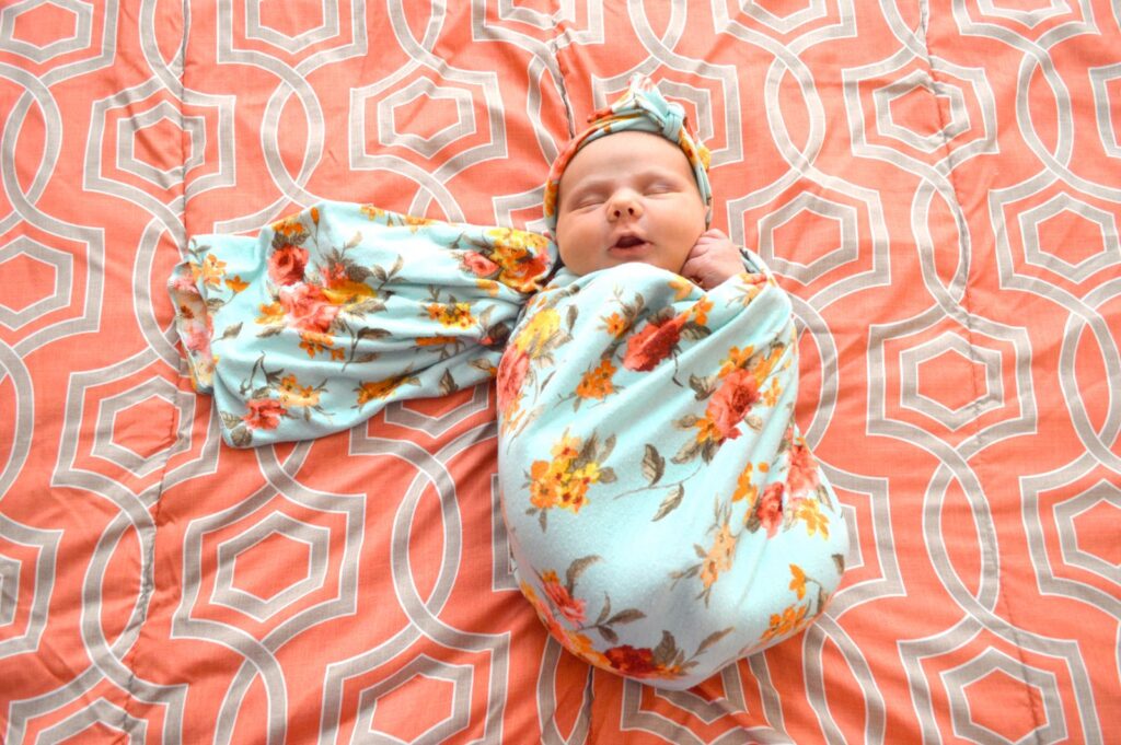 Newborn wrapped in a floral muslin swaddle on a coral blanket