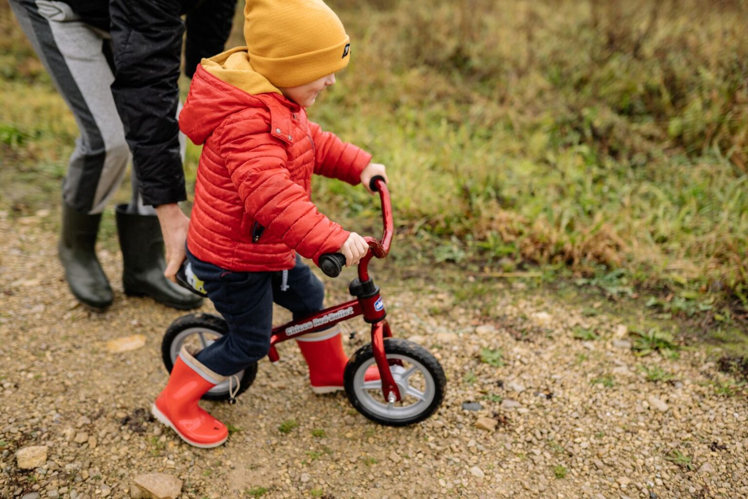 Toddler in a red jacket riding a red balance bike with a parent walking alongside